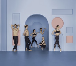 A group of dancers is positioned in a minimalistic setting with geometric shapes on the walls. The dancers are dressed in black dance apparel, with some performing ballet poses and others resting. The setting features a color scheme of blue and pink.