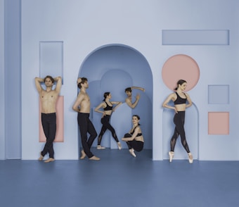 A group of dancers is positioned in a minimalistic setting with geometric shapes on the walls. The dancers are dressed in black dance apparel, with some performing ballet poses and others resting. The setting features a color scheme of blue and pink.
