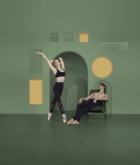 A ballet dancer poses gracefully on pointe in front of a minimalist backdrop, which features geometric cutouts in yellow and green. To the side, a shirtless man reclines in a chair, observing the dancer.