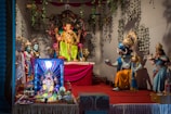 A vibrant and intricately decorated scene featuring several Hindu deities represented as statues, with the central focus on a large Ganesha idol adorned with colorful garments and jewelry. Surrounding Ganesha are various other statues including what appears to be a deity with a boar's head holding a globe, and another deity playing a musical instrument. The setting is adorned with green vines and is brightly lit.
