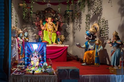 A vibrant and intricately decorated scene featuring several Hindu deities represented as statues, with the central focus on a large Ganesha idol adorned with colorful garments and jewelry. Surrounding Ganesha are various other statues including what appears to be a deity with a boar's head holding a globe, and another deity playing a musical instrument. The setting is adorned with green vines and is brightly lit.