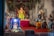 A vibrant and intricately decorated scene featuring several Hindu deities represented as statues, with the central focus on a large Ganesha idol adorned with colorful garments and jewelry. Surrounding Ganesha are various other statues including what appears to be a deity with a boar's head holding a globe, and another deity playing a musical instrument. The setting is adorned with green vines and is brightly lit.