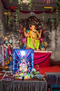 A colorful altar featuring statues of Hindu deities, with a prominent figure of Ganesha adorned in bright clothing and jewelry. The altar is decorated with garlands and various offerings, set against a backdrop of greenery and red drapery.