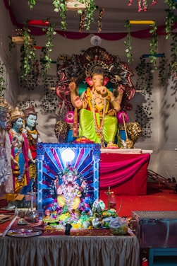 A colorful altar featuring statues of Hindu deities, with a prominent figure of Ganesha adorned in bright clothing and jewelry. The altar is decorated with garlands and various offerings, set against a backdrop of greenery and red drapery.
