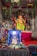 A colorful altar featuring statues of Hindu deities, with a prominent figure of Ganesha adorned in bright clothing and jewelry. The altar is decorated with garlands and various offerings, set against a backdrop of greenery and red drapery.