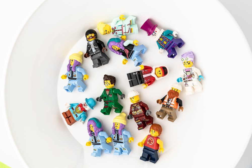 A colorful assortment of LEGO minifigures scattered on a white surface, displaying a variety of characters including a figure in a black ninja outfit, one wearing a purple top hat, and another dressed as a clown
