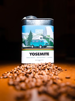 A metal canister labeled 'Yosemite' with a design featuring a blue van and a mountainous landscape, likely representing a coffee product. In front, there is a pile of coffee beans scattered on a wooden surface.
