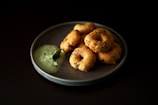 A plate of vadai, a type of savory Indian fritter, arranged neatly with a serving of green chutney on the side. The vadai are golden brown with visible herbs and spices, and the chutney appears creamy with a leaf garnish.