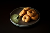 A plate of vadai, a type of savory Indian fritter, arranged neatly with a serving of green chutney on the side. The vadai are golden brown with visible herbs and spices, and the chutney appears creamy with a leaf garnish.
