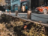 A street scene featuring a building entrance with two people working, possibly moving a large item wrapped in a yellow covering. There is a red-orange car parked nearby. The area in front of the building has a sidewalk bordered by stone pillars connected by chains. The foreground has a small garden with plants and flowers.