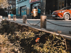 A street scene featuring a building entrance with two people working, possibly moving a large item wrapped in a yellow covering. There is a red-orange car parked nearby. The area in front of the building has a sidewalk bordered by stone pillars connected by chains. The foreground has a small garden with plants and flowers.