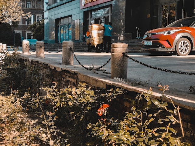 A street scene featuring a building entrance with two people working, possibly moving a large item wrapped in a yellow covering. There is a red-orange car parked nearby. The area in front of the building has a sidewalk bordered by stone pillars connected by chains. The foreground has a small garden with plants and flowers.