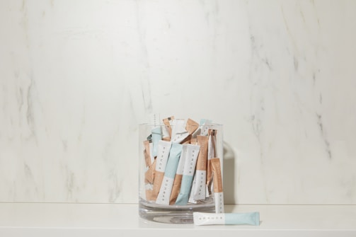 A glass container is filled with several sachets labeled 'SPRUCE' in a minimalist style, placed against a marble-textured backdrop. The sachets are predominantly blue and beige.
