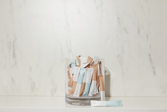 A glass container is filled with several sachets labeled 'SPRUCE' in a minimalist style, placed against a marble-textured backdrop. The sachets are predominantly blue and beige.