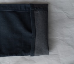 A close-up view of the hem of a pair of dark denim jeans, showcasing the fabric texture and stitching. The jeans are folded with the interior and exterior sides visible against a light, textured background.