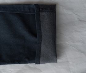 A close-up view of the hem of a pair of dark denim jeans, showcasing the fabric texture and stitching. The jeans are folded with the interior and exterior sides visible against a light, textured background.