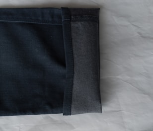 A close-up view of the hem of a pair of dark denim jeans, showcasing the fabric texture and stitching. The jeans are folded with the interior and exterior sides visible against a light, textured background.