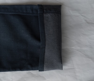 A close-up view of the hem of a pair of dark denim jeans, showcasing the fabric texture and stitching. The jeans are folded with the interior and exterior sides visible against a light, textured background.