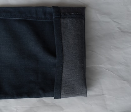 A close-up view of the hem of a pair of dark denim jeans, showcasing the fabric texture and stitching. The jeans are folded with the interior and exterior sides visible against a light, textured background.