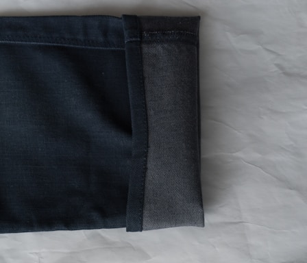 A close-up view of the hem of a pair of dark denim jeans, showcasing the fabric texture and stitching. The jeans are folded with the interior and exterior sides visible against a light, textured background.