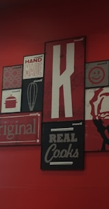 A collection of vintage-style wall art with various designs, including a large letter 'K', the words 'HAND MADE', 'REAL Cooks', 'original', and a whisk illustration. The background is a deep red, with the artwork featuring a mix of black, white, and red tones.