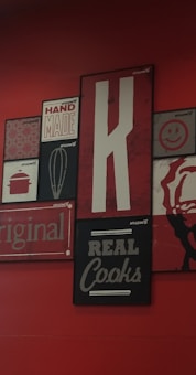 A collection of vintage-style wall art with various designs, including a large letter 'K', the words 'HAND MADE', 'REAL Cooks', 'original', and a whisk illustration. The background is a deep red, with the artwork featuring a mix of black, white, and red tones.