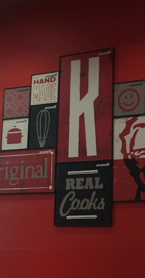 A collection of vintage-style wall art with various designs, including a large letter 'K', the words 'HAND MADE', 'REAL Cooks', 'original', and a whisk illustration. The background is a deep red, with the artwork featuring a mix of black, white, and red tones.