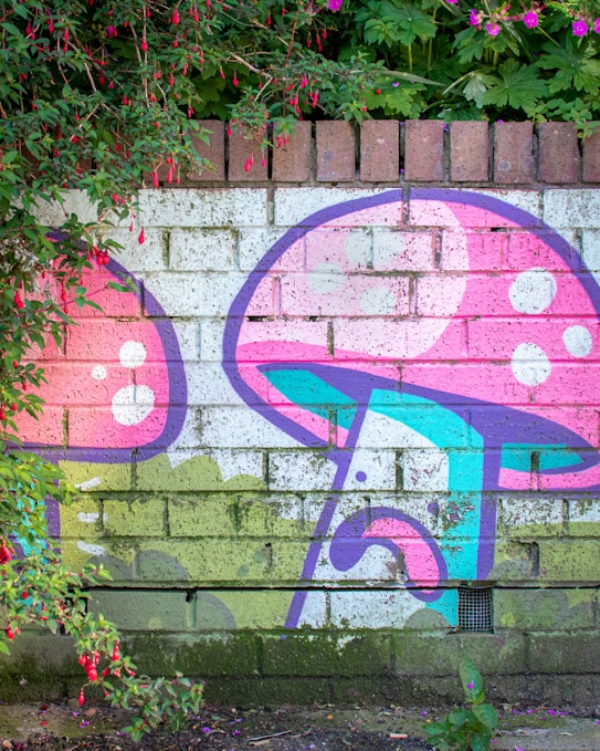 A vibrant piece of street art featuring colorful mushrooms painted on a brick wall. The artwork incorporates pink, purple, blue, and white colors, with lush greenery and red flowers hanging around the mural. The background is composed of unpainted bricks, adding texture to the scene.