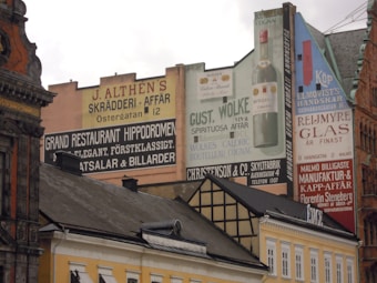 An urban scene featuring a row of historic buildings adorned with large, colorful advertisements painted on their facades. The ads promote various businesses, including a restaurant, a tailor's shop, a handkerchief store, and a spirits retailer. The architecture reflects a mix of classic styles with ornate details and sloped roofs.
