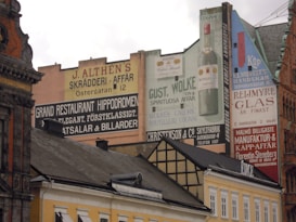 An urban scene featuring a row of historic buildings adorned with large, colorful advertisements painted on their facades. The ads promote various businesses, including a restaurant, a tailor's shop, a handkerchief store, and a spirits retailer. The architecture reflects a mix of classic styles with ornate details and sloped roofs.