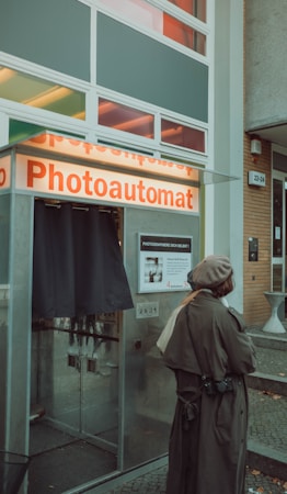 A person wearing a long coat and beret stands in front of a photo booth labeled 'Photoautomat.' The booth is situated outside a building with large windows and a panel displaying instructions.
