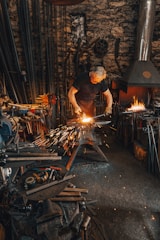 A person is working in a blacksmith's workshop, hammering a metal piece on an anvil with sparks flying. The workshop is filled with tools, equipment, and a lit furnace in the background. The walls are made of stone and the atmosphere is gritty and industrious.