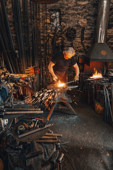 A person is working in a blacksmith's workshop, hammering a metal piece on an anvil with sparks flying. The workshop is filled with tools, equipment, and a lit furnace in the background. The walls are made of stone and the atmosphere is gritty and industrious.