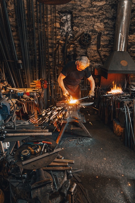 A person is working in a blacksmith's workshop, hammering a metal piece on an anvil with sparks flying. The workshop is filled with tools, equipment, and a lit furnace in the background. The walls are made of stone and the atmosphere is gritty and industrious.