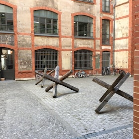 A brick building with arched windows and barred grilles surrounds a cobblestone courtyard. Three wooden anti-tank obstacles are placed in the middle of the courtyard. Several bicycles are parked against the building, adding a touch of everyday life to the scene.