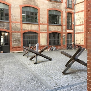 A brick building with arched windows and barred grilles surrounds a cobblestone courtyard. Three wooden anti-tank obstacles are placed in the middle of the courtyard. Several bicycles are parked against the building, adding a touch of everyday life to the scene.