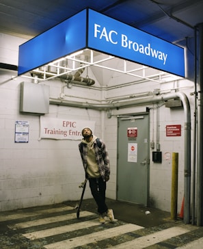 A person holding a skateboard stands in an indoor setting, beneath a blue sign that reads 'FAC Broadway'. The setting appears to be a parking garage or an industrial area, with exposed pipes and signs indicating an EPIC Training Entrance. The person is wearing casual clothing with a plaid shirt and beanie.