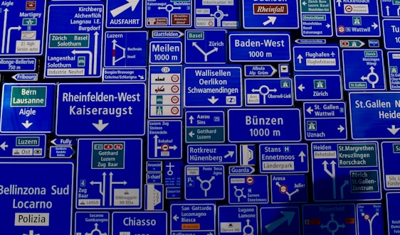 A collage of numerous European road signs displaying various destinations, directions, distances, and traffic symbols. The signs are primarily blue with white text and symbols, intermixed with some green signs. They feature names of cities, highways, and exits.
