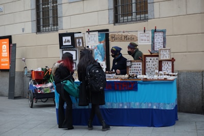 A small street market stall selling handmade jewelry and crafts, with two vendors behind the table and two customers looking at the items. The stall is decorated with frames and signs, including one that reads 'Hand Made.' The atmosphere is casual with a blue tablecloth covering the display.
