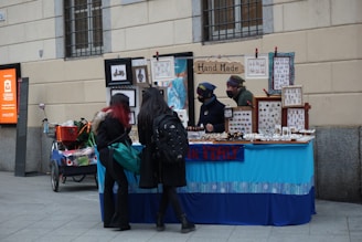 A small street market stall selling handmade jewelry and crafts, with two vendors behind the table and two customers looking at the items. The stall is decorated with frames and signs, including one that reads 'Hand Made.' The atmosphere is casual with a blue tablecloth covering the display.