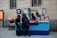 A small street market stall selling handmade jewelry and crafts, with two vendors behind the table and two customers looking at the items. The stall is decorated with frames and signs, including one that reads 'Hand Made.' The atmosphere is casual with a blue tablecloth covering the display.