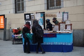 A small street market stall selling handmade jewelry and crafts, with two vendors behind the table and two customers looking at the items. The stall is decorated with frames and signs, including one that reads 'Hand Made.' The atmosphere is casual with a blue tablecloth covering the display.