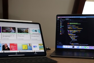 A workspace featuring a tablet and a laptop on a wooden desk. The tablet displays an online learning platform with various course thumbnails, while the laptop shows a coding environment with a colorful code editor. The setup suggests a focus on learning and coding.