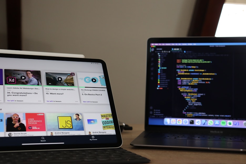 A workspace featuring a tablet and a laptop on a wooden desk. The tablet displays an online learning platform with various course thumbnails, while the laptop shows a coding environment with a colorful code editor. The setup suggests a focus on learning and coding.