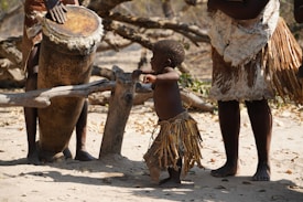 A young child wearing traditional attire with natural materials is curiously interacting with a large drum being played by an adult. The scene seems to be part of a cultural or traditional event, with people wearing garments made from natural fibers. The ground is sandy, and there are wooden logs and branches around.