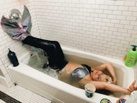 A person is lying in a bathtub wearing a colorful mermaid costume with a shimmering, scaly tail. The bathtub is filled with water and the surrounding area has white tiled walls. There are various objects around the bathtub, including a green bottle of soap, a black bottle, and a cup with a spoon in it. The person appears to be content or relaxed.