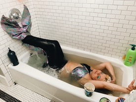 A person is lying in a bathtub wearing a colorful mermaid costume with a shimmering, scaly tail. The bathtub is filled with water and the surrounding area has white tiled walls. There are various objects around the bathtub, including a green bottle of soap, a black bottle, and a cup with a spoon in it. The person appears to be content or relaxed.