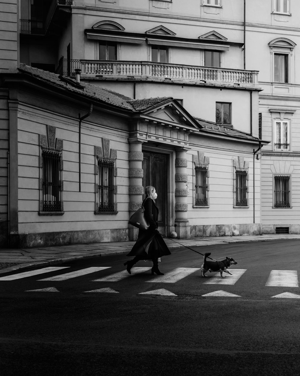 grayscale photo of woman walking on sidewalk near building