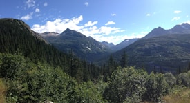 A vast and scenic mountain landscape featuring towering peaks under a clear blue sky with scattered clouds. Dense forests and lush greenery cover the slopes and valleys, creating a serene and natural setting.
