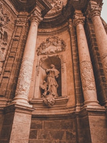 A detailed stone sculpture depicting a robed figure standing in a niche, surrounded by ornate carvings and flanked by grand columns. The rich textures of the stone and intricate details of the artwork are highlighted, creating a sense of historical and architectural grandeur.